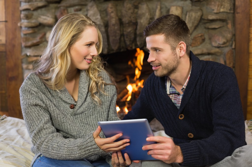 Happy lovely young couple using tablet PC in front of lit fireplace